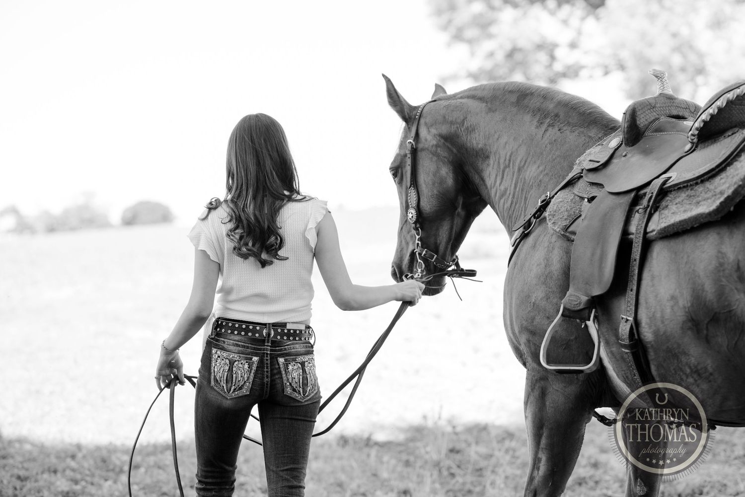 Black and white image of a woman leading a saddled horse through a field.