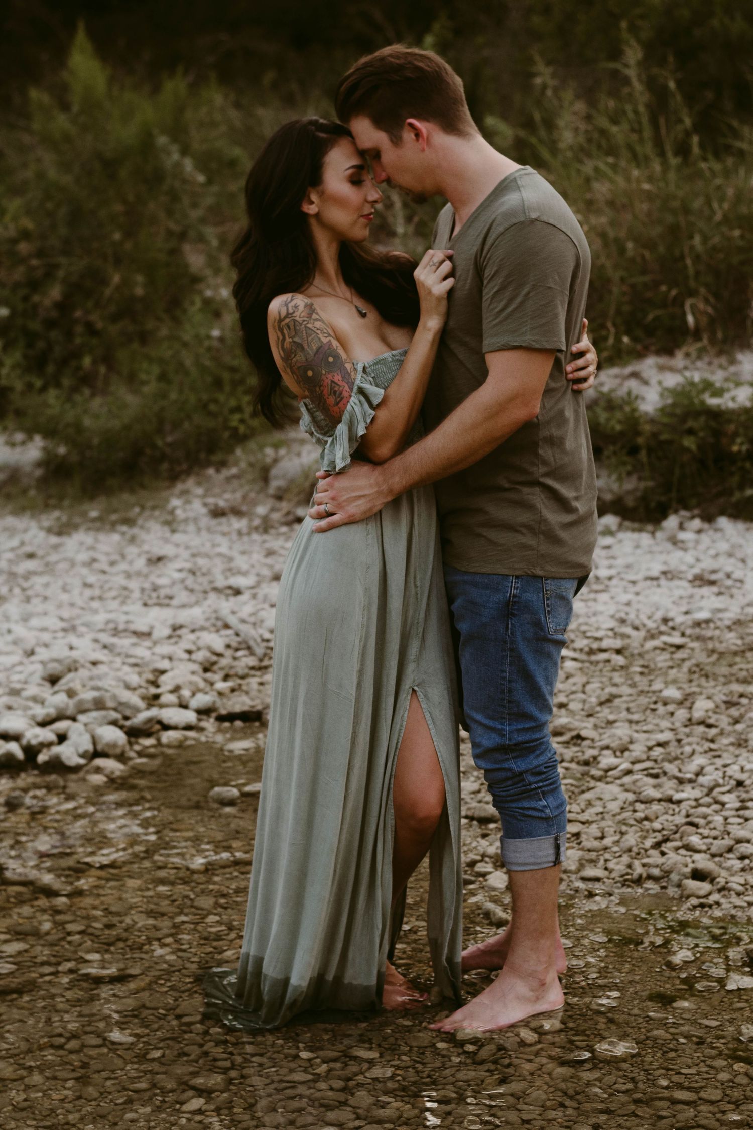 Romantic couple embraces in a natural setting wearing casual clothes near a rocky riverbed at sunset.