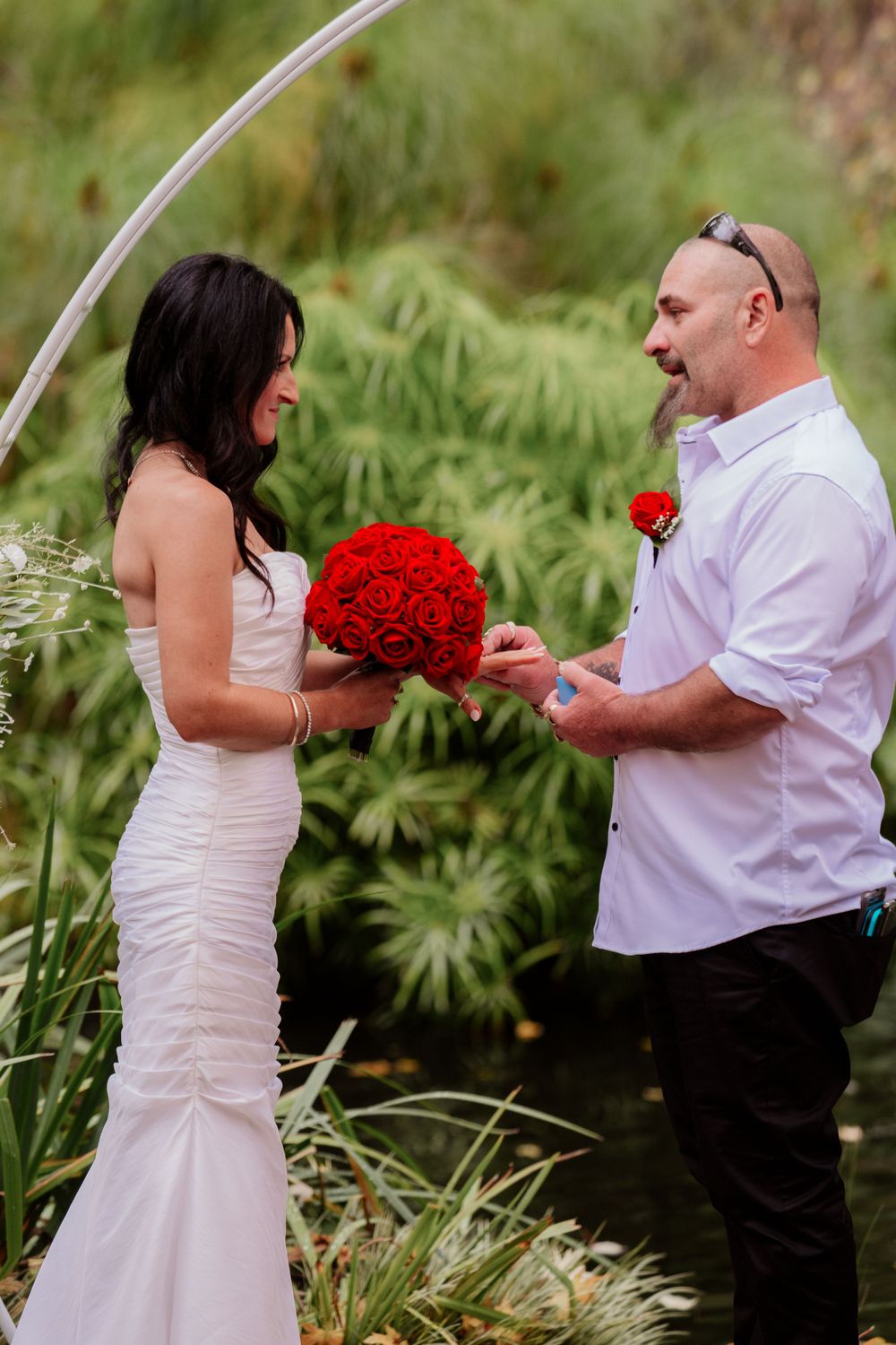 Wedding photoshoot in garden setting with white dress and vibrant red rose bouquet.