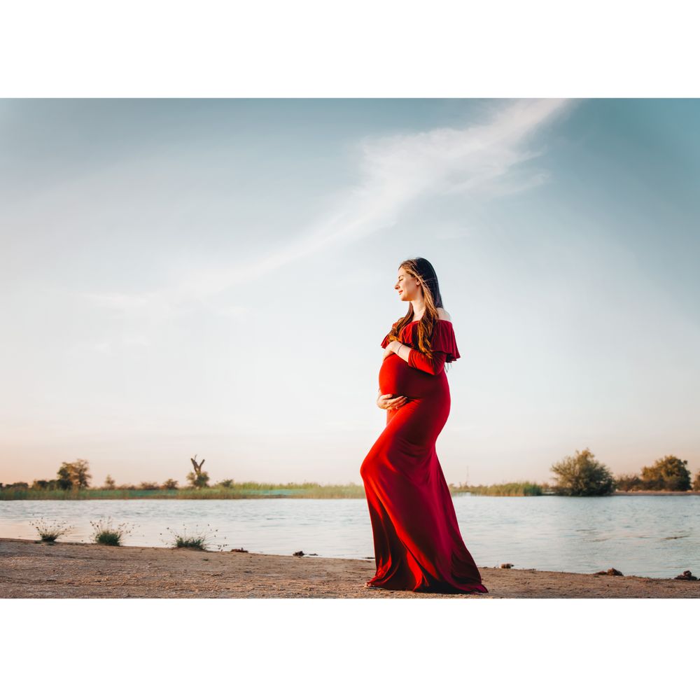 Pregnant lady in deep red gown standing by a lake during golden hour.