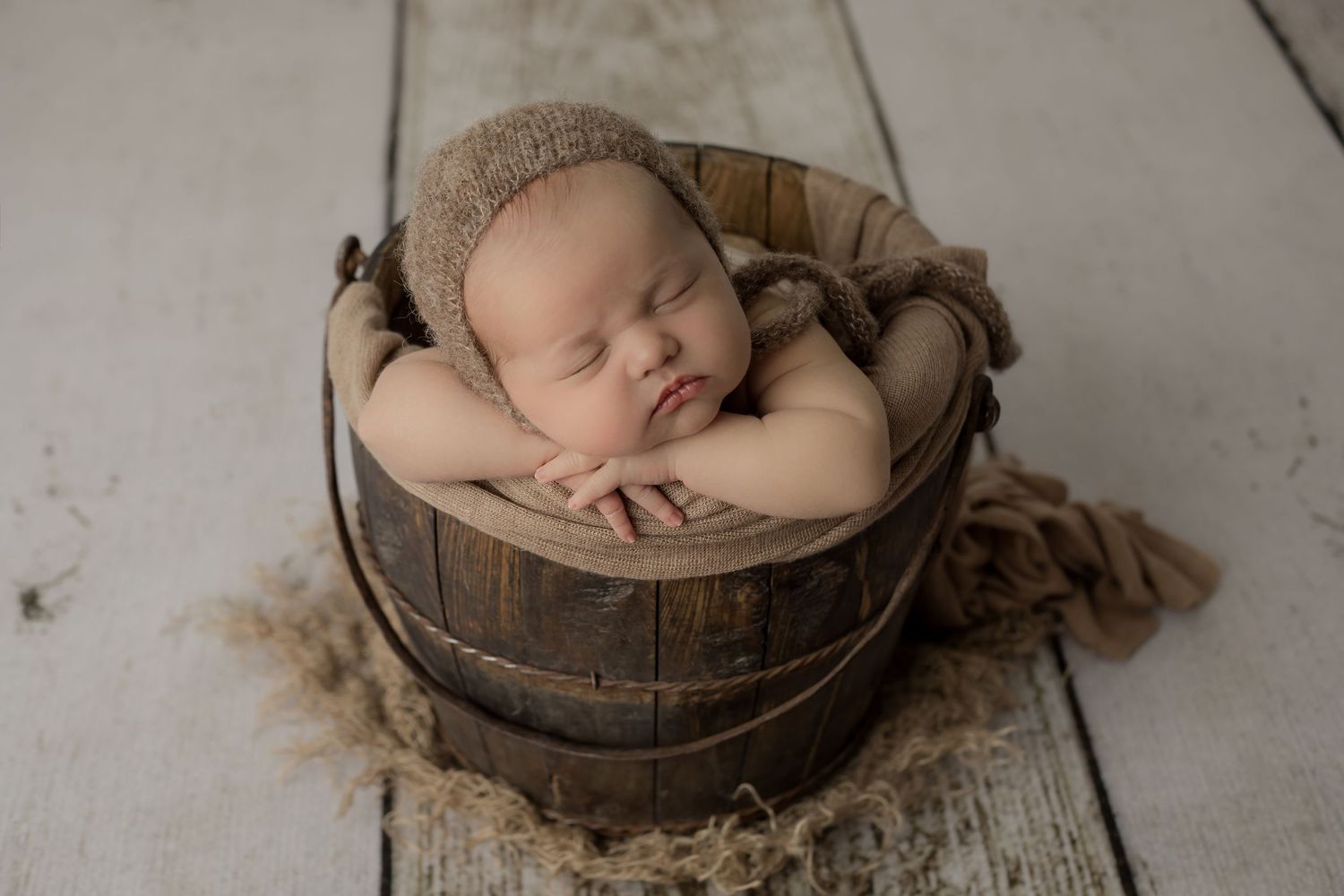 Baby sleeping boy with chubby cheeks safely posed in a brown bucket with a handsome bonnet on for his newborn studio portrait session.