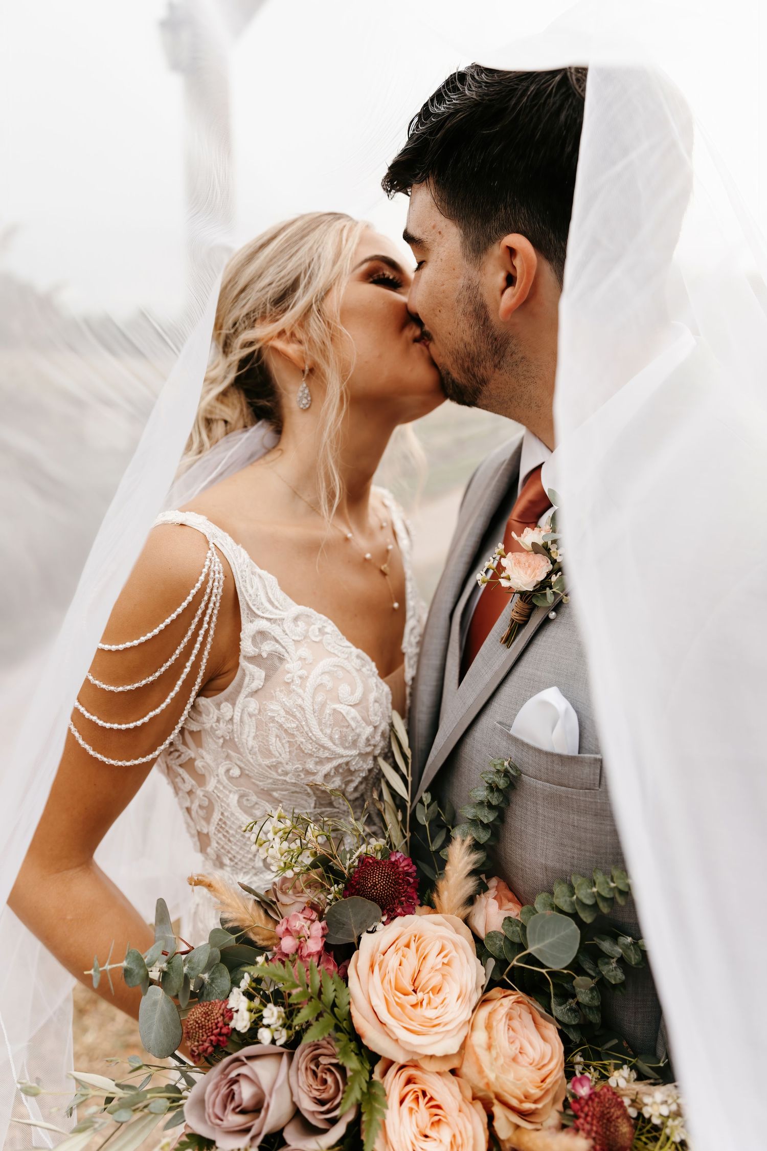 Bride and groom kissing under a flowing veil, bride holding a lush floral bouquet.