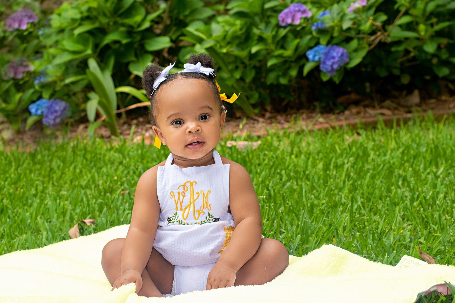 Baby in white outfit sitting on yellow blanket with flower garden background.