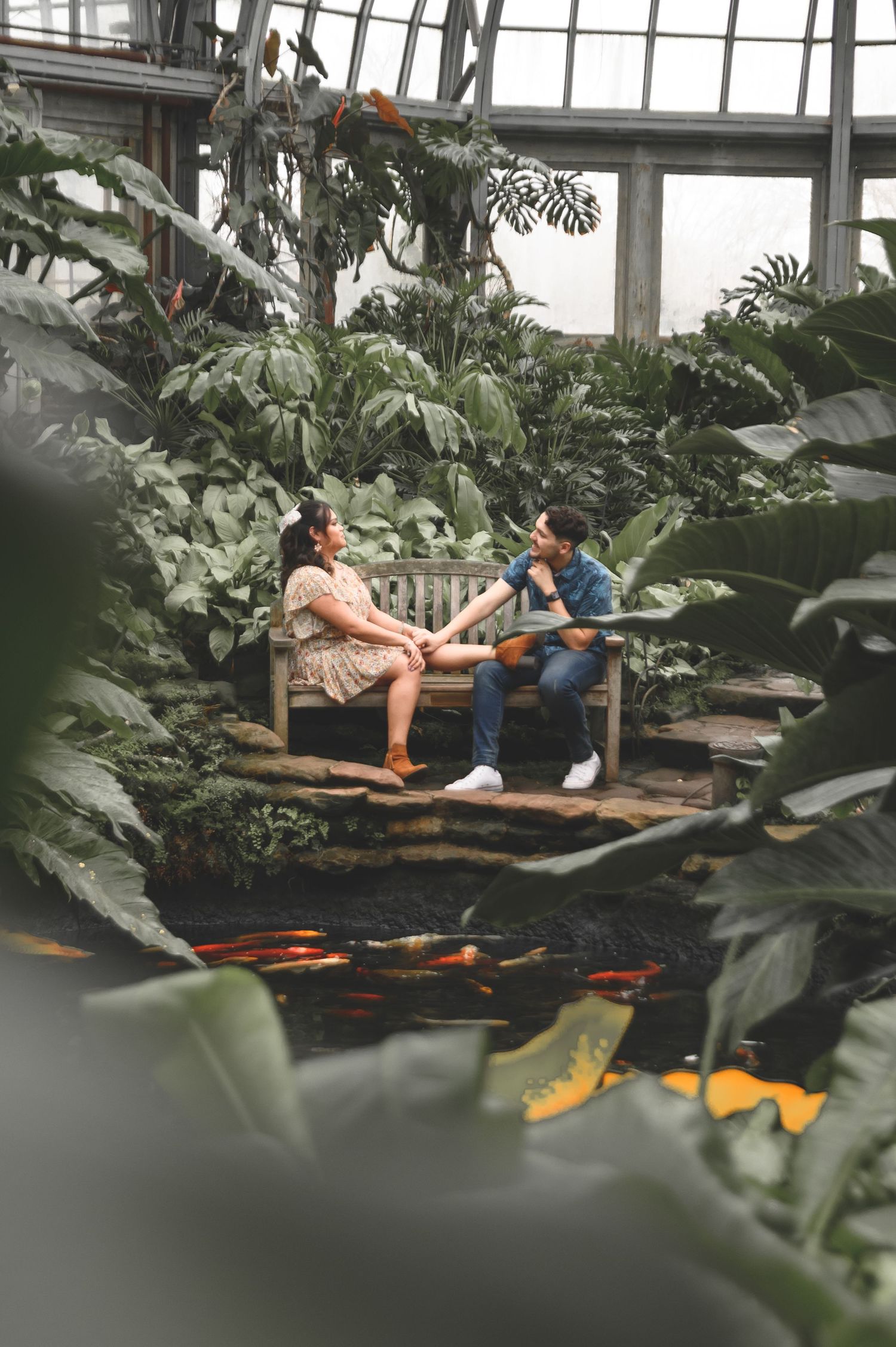 A couple enjoys a romantic moment surrounded by lush tropical plants in a greenhouse setting.