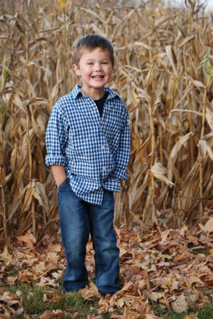 Child wearing blue checkered shirt and jeans stands in front of dried corn stalks on an autumn day.