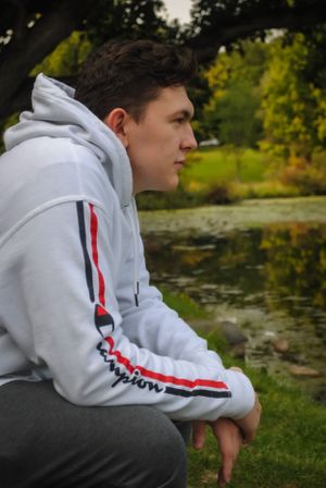 Profile view of person in white athletic hoodie sitting near pond in park setting.