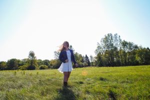 Woman in white dress with backpack standing in sunlit meadow during golden hour.