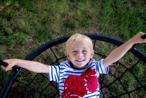 Child in striped shirt with red design spreads arms while sitting on playground equipment.