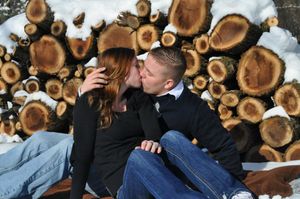 A couple shares an intimate moment while sitting on stacked logs in winter.