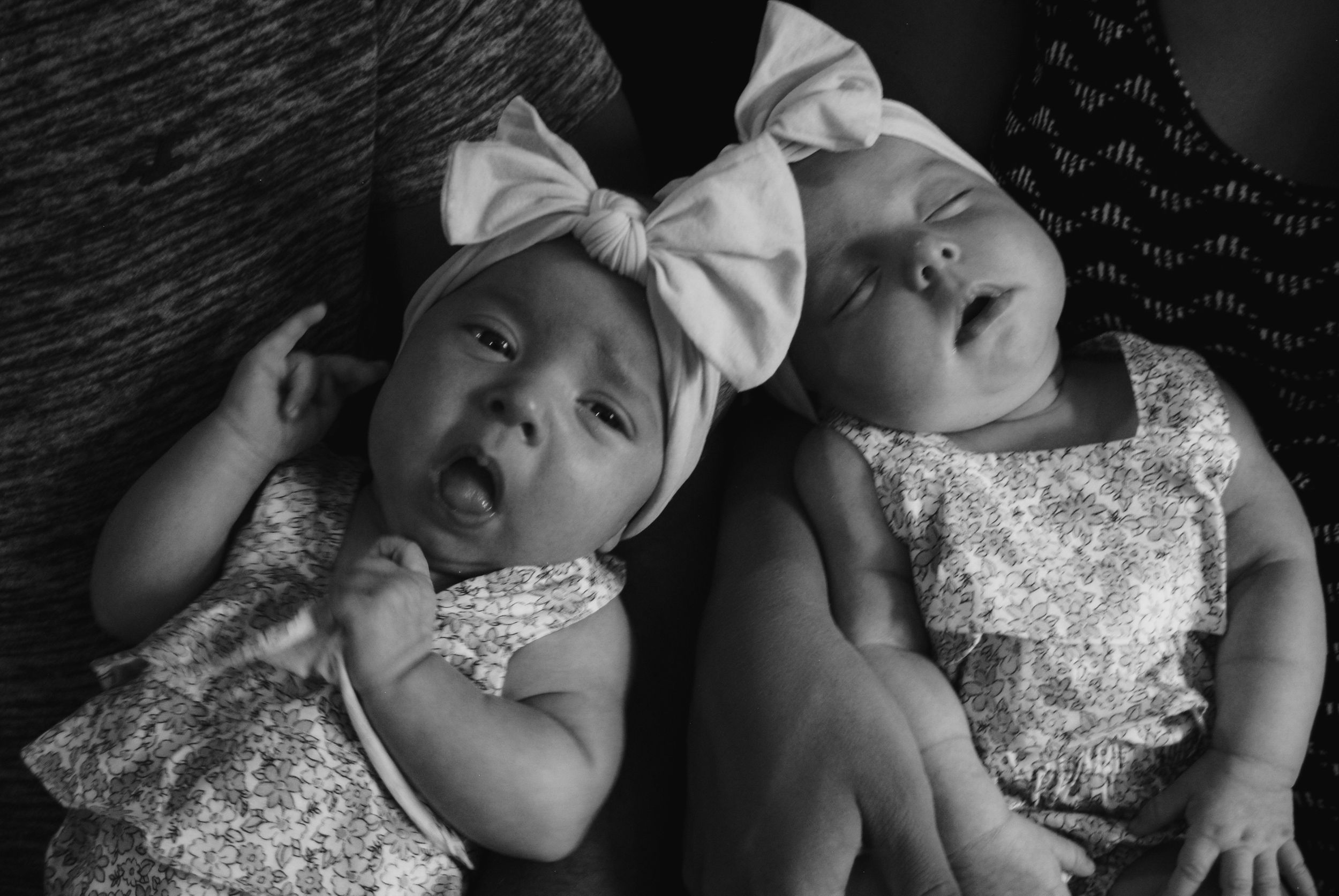 Two babies wearing matching white dresses and bows rest together in black and white portrait.