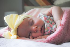 Sleeping baby with yellow bow wearing floral dress wrapped in pink blanket.