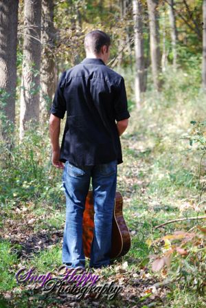 Person in dark shirt holding guitar while standing in wooded area.