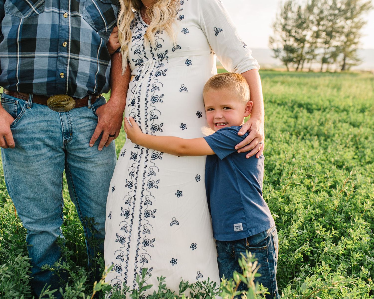 A small child in denim hugs a parent wearing a floral dress in a green field.