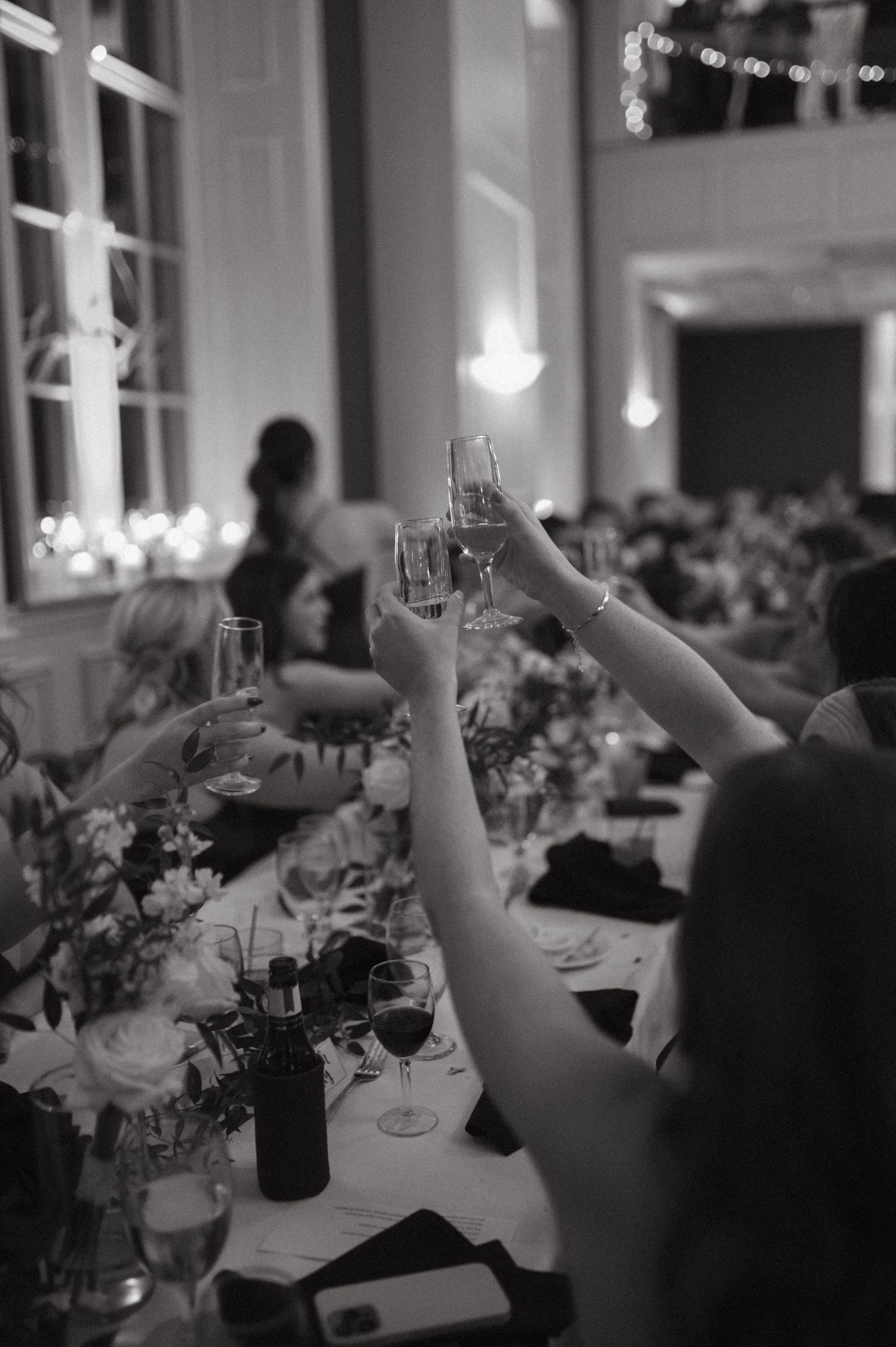 Black and white photo of wedding guests raising glasses in a toast during reception dinner.