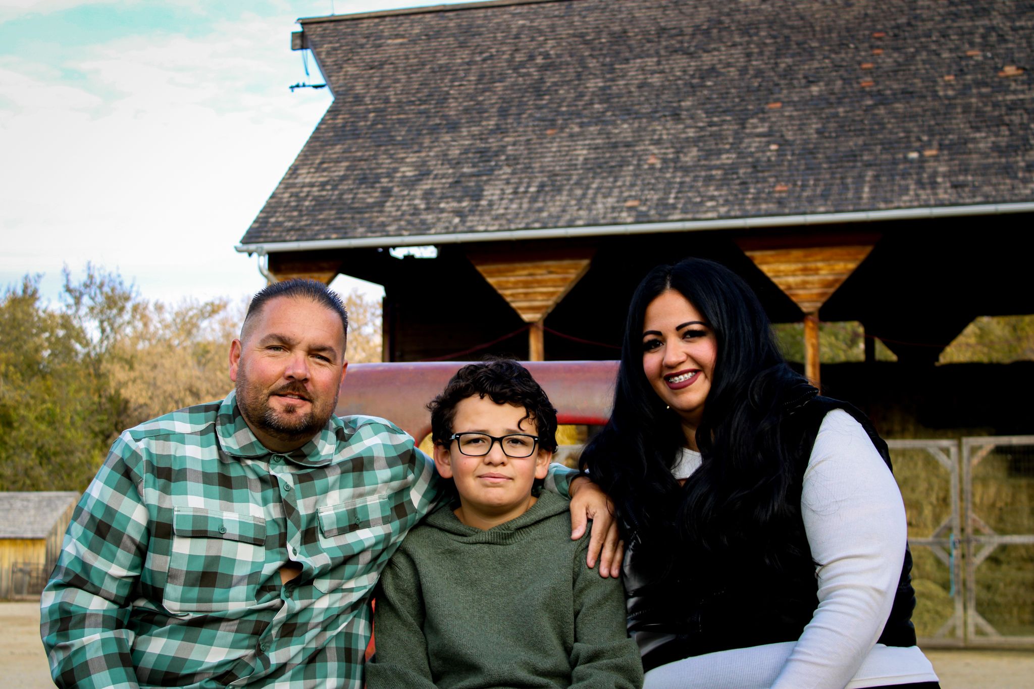 A family of three poses together in front of a wooden shelter during fall.