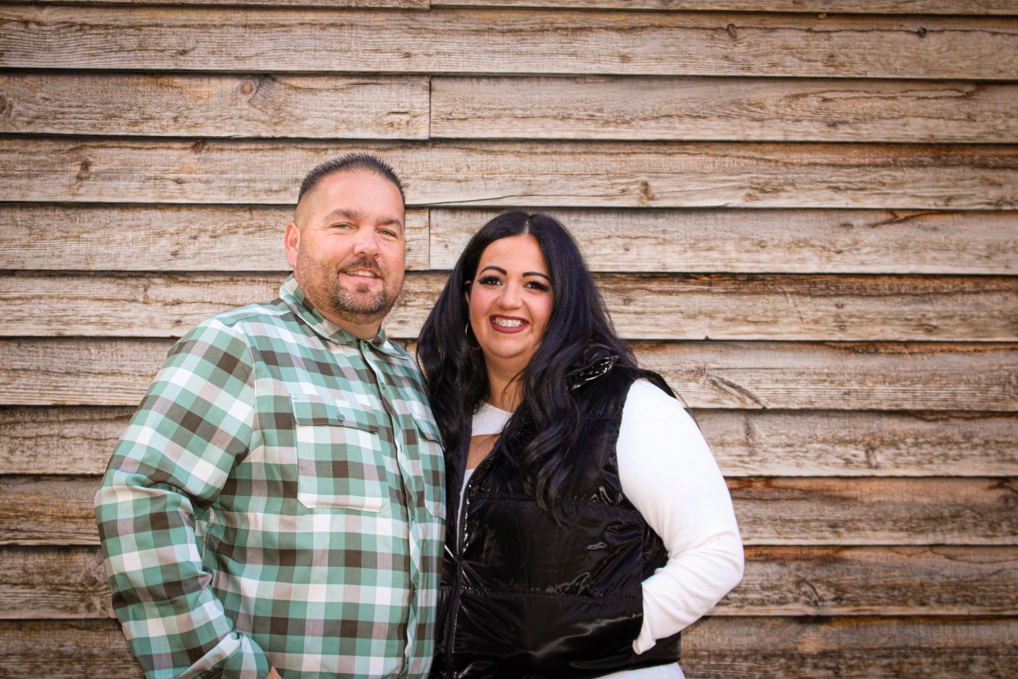 Two people smile together against rustic wooden wall background.