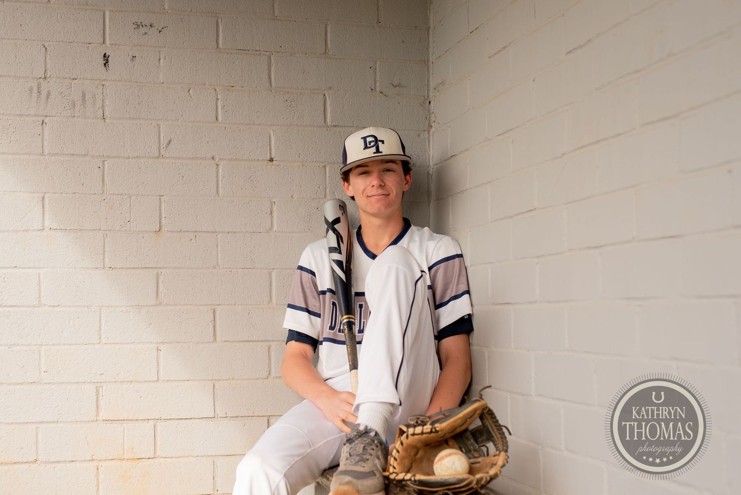 Baseball player in white uniform sits casually in dugout with baseball glove and navy cap.