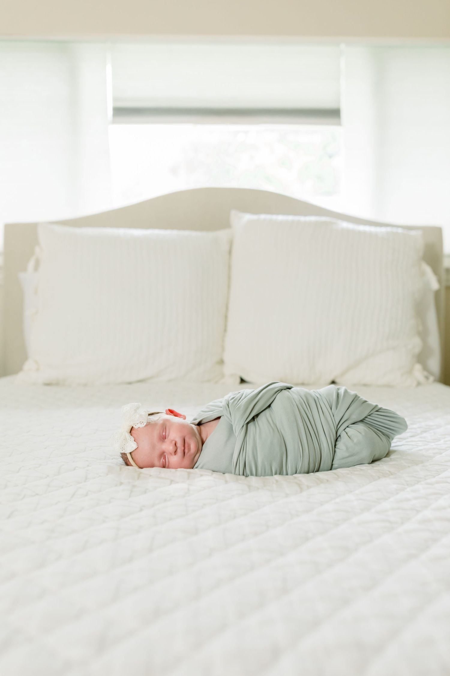 A swaddled newborn in sage green sleeps peacefully on a white bed.