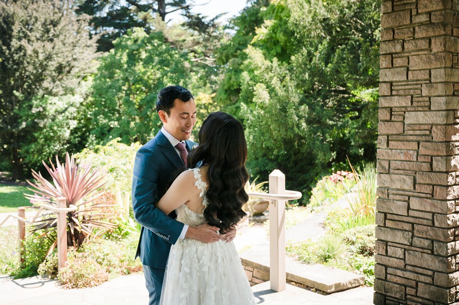 A couple in wedding attire embraces under towering trees and lush greenery.