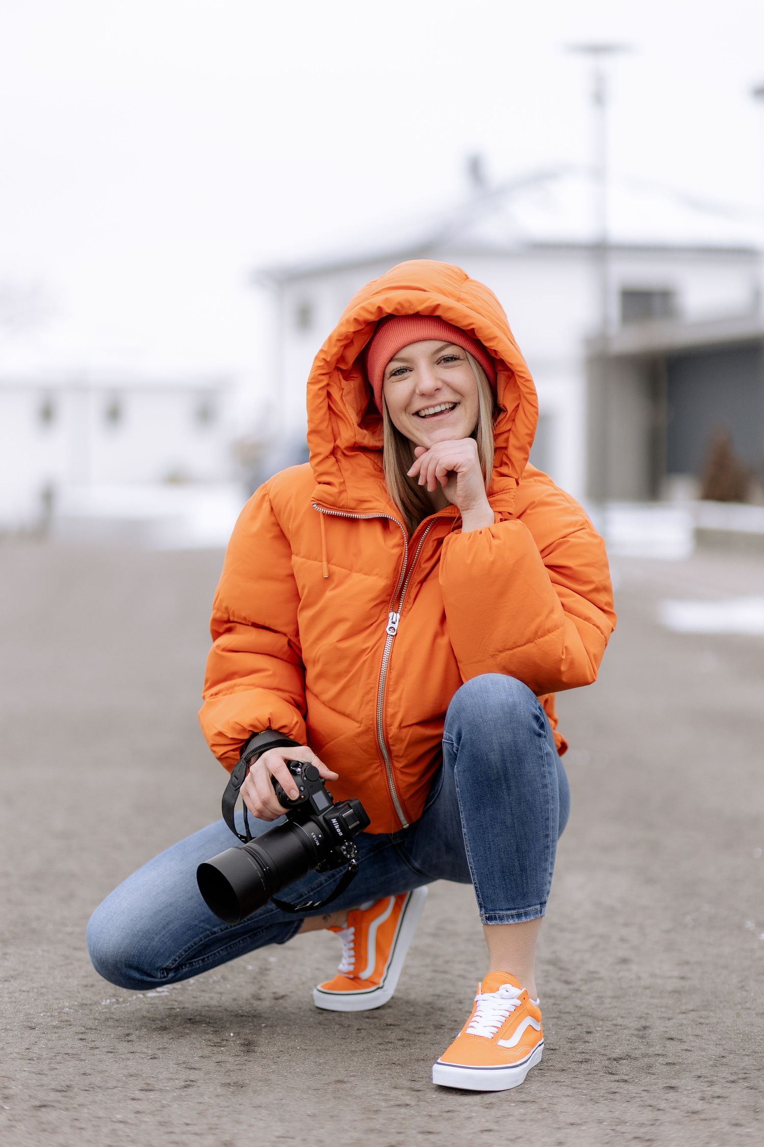 Eine Person in einer leuchtend orangefarbenen Daunenjacke und Jeans hockt sich hin, während sie weiße und orangefarbene Turnschuhe trägt.
