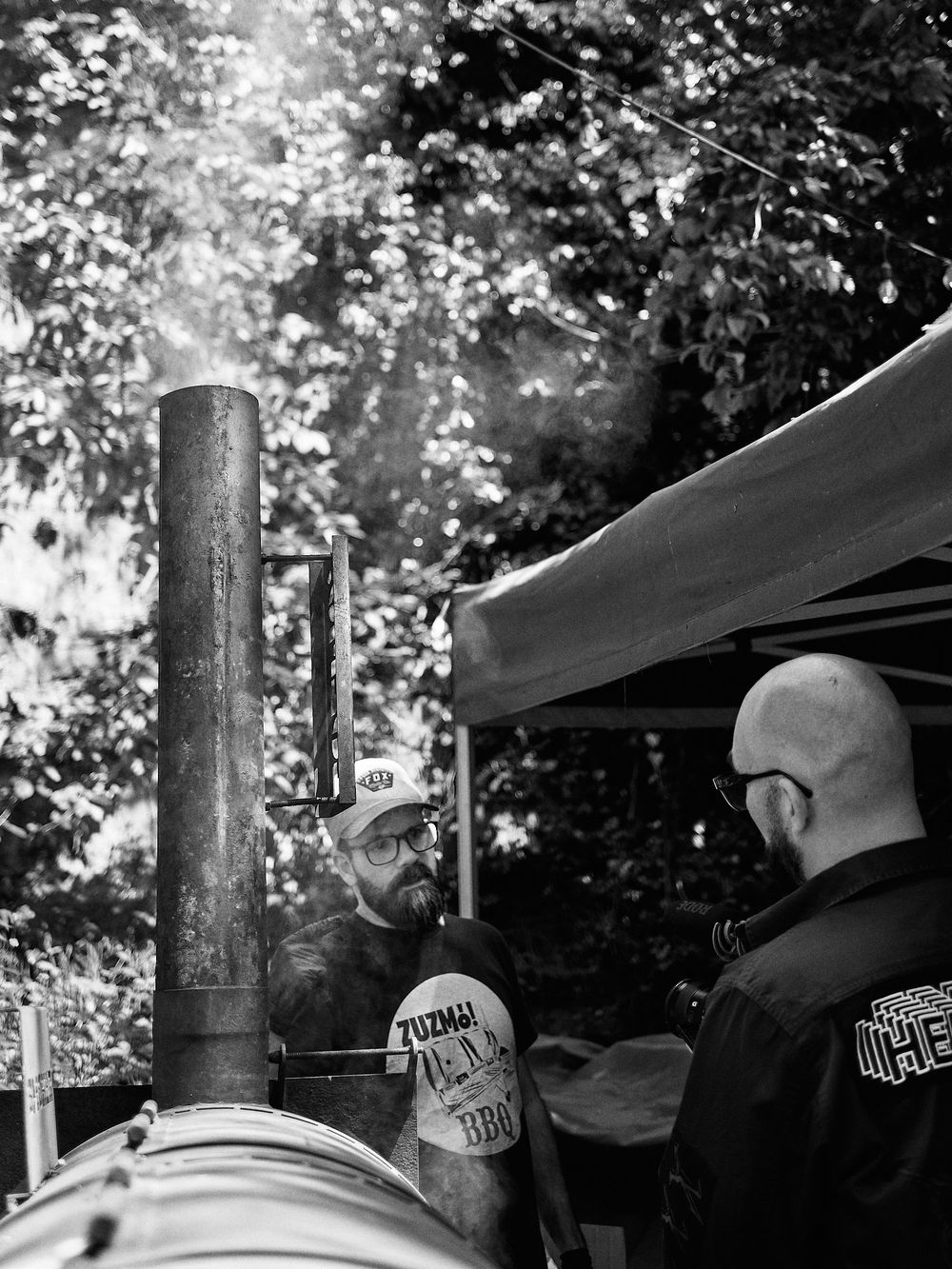 Two people stand by a large smoker setup discussing barbecue techniques outdoors.