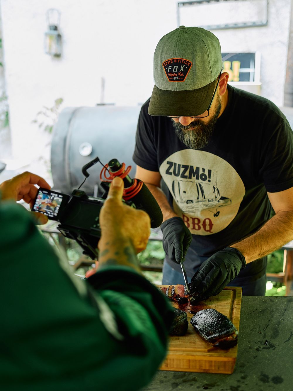 BBQ preparation scene with cook wearing Zuzma BBQ shirt and green cap.