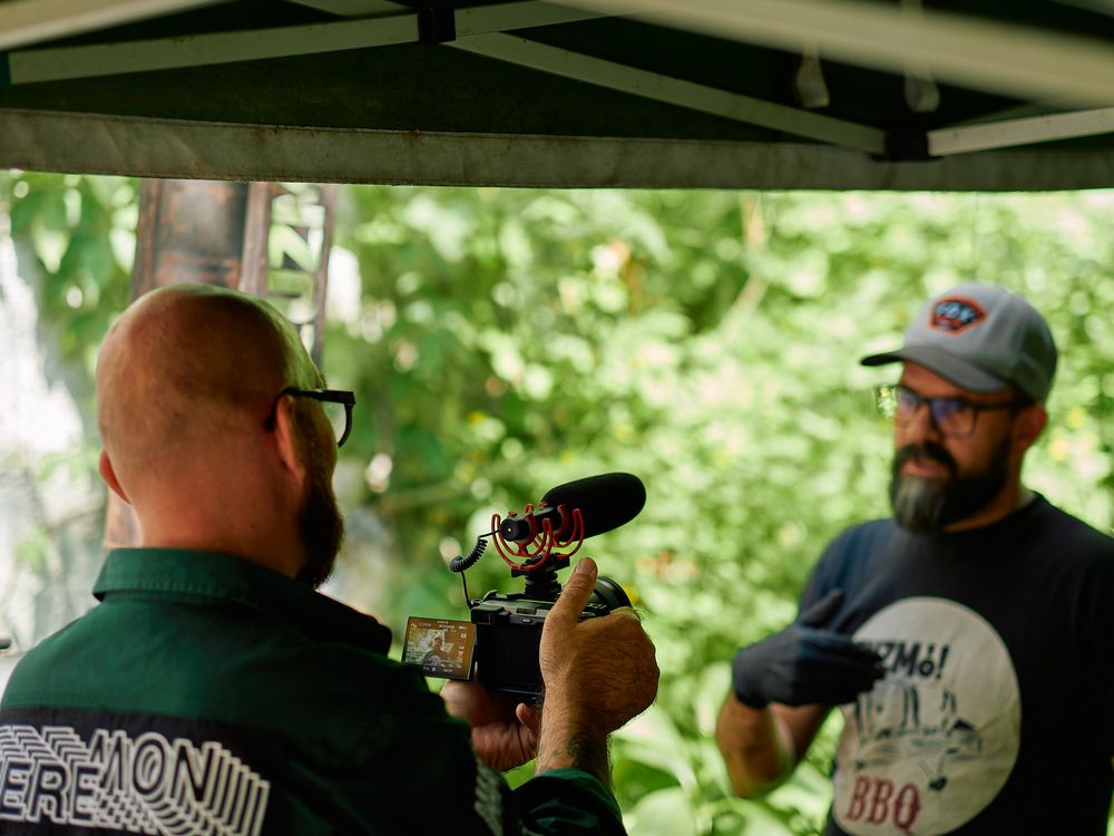 Camera operators filming BBQ preparation under a white tent with natural greenery background.
