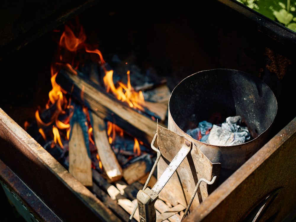 A camp fire burns brightly with wooden logs while charcoal smolders in a metal container nearby.