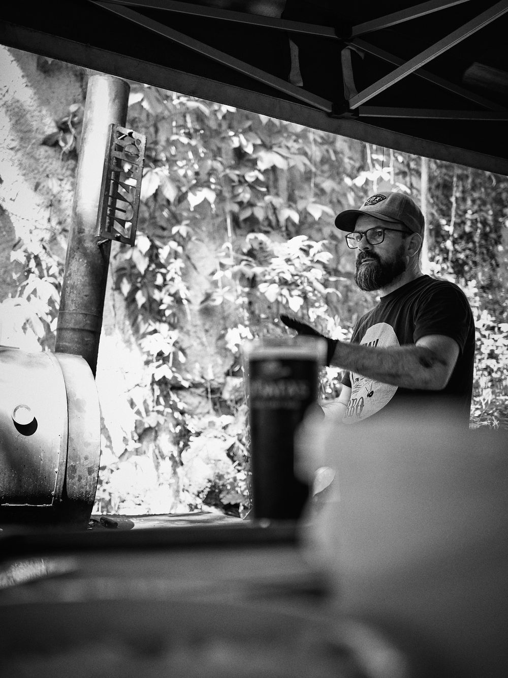 Black and white photo of BBQ smoker operation under outdoor canopy.