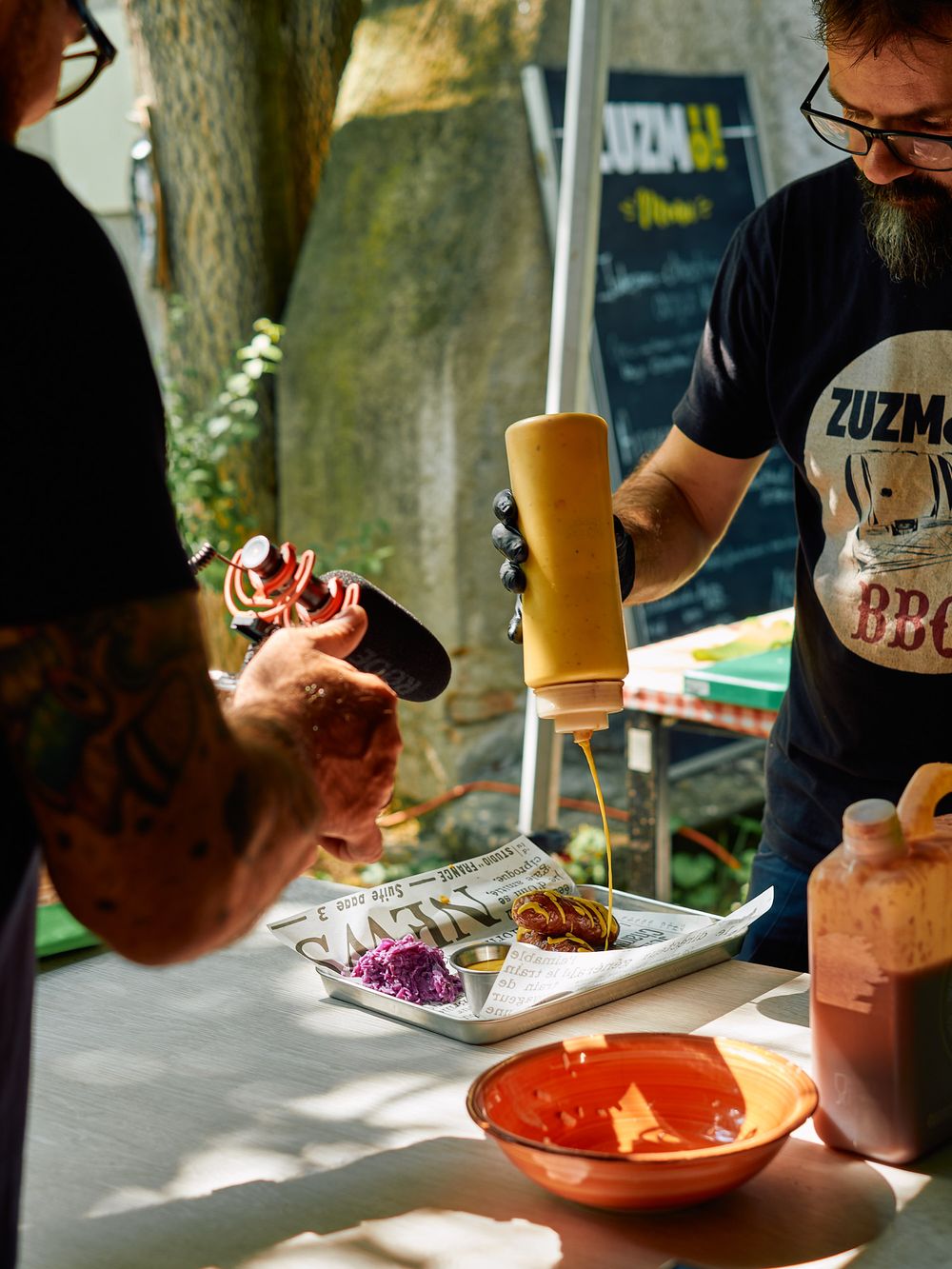 Food preparation scene with plates and dishes being arranged on a white tablecloth outdoors.