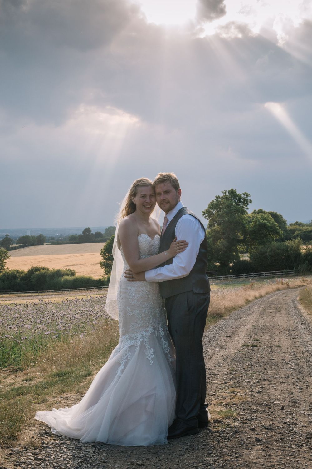 A romantic wedding couple embraces on a country path with dramatic cloudy skies in the background.