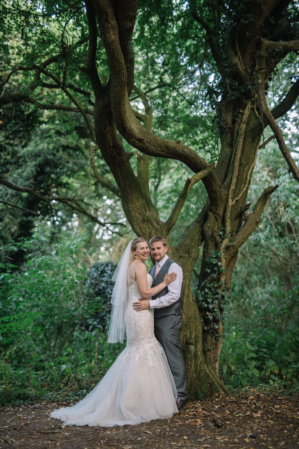 A couple in wedding attire poses together against a twisted tree trunk in a lush forest setting.