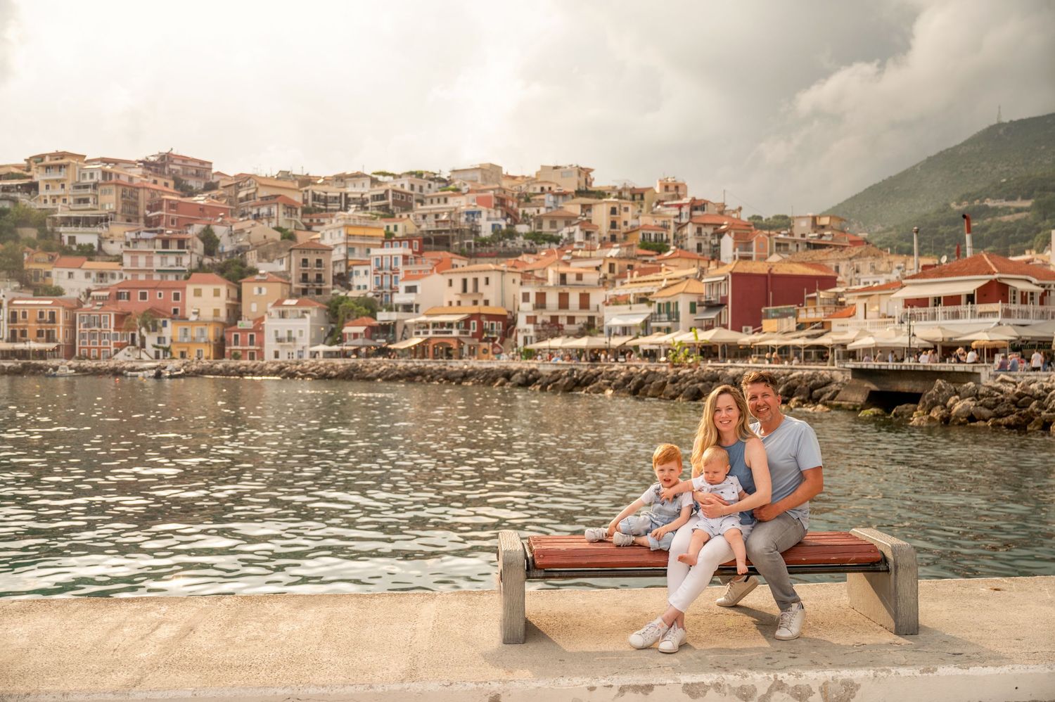 Young couple with children sitting on harbor bench overlooking coastal village with colorful buildings.
