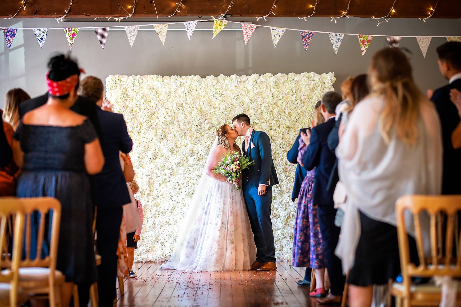 Bride and groom kiss standing before a white flower wall during their wedding at The Castlefield Rooms in Manchester