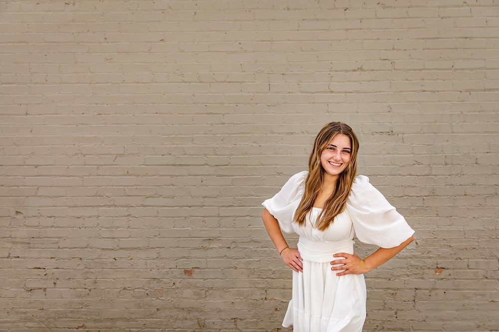 senior photo session of girl in white dress standing in front of painted brick wall