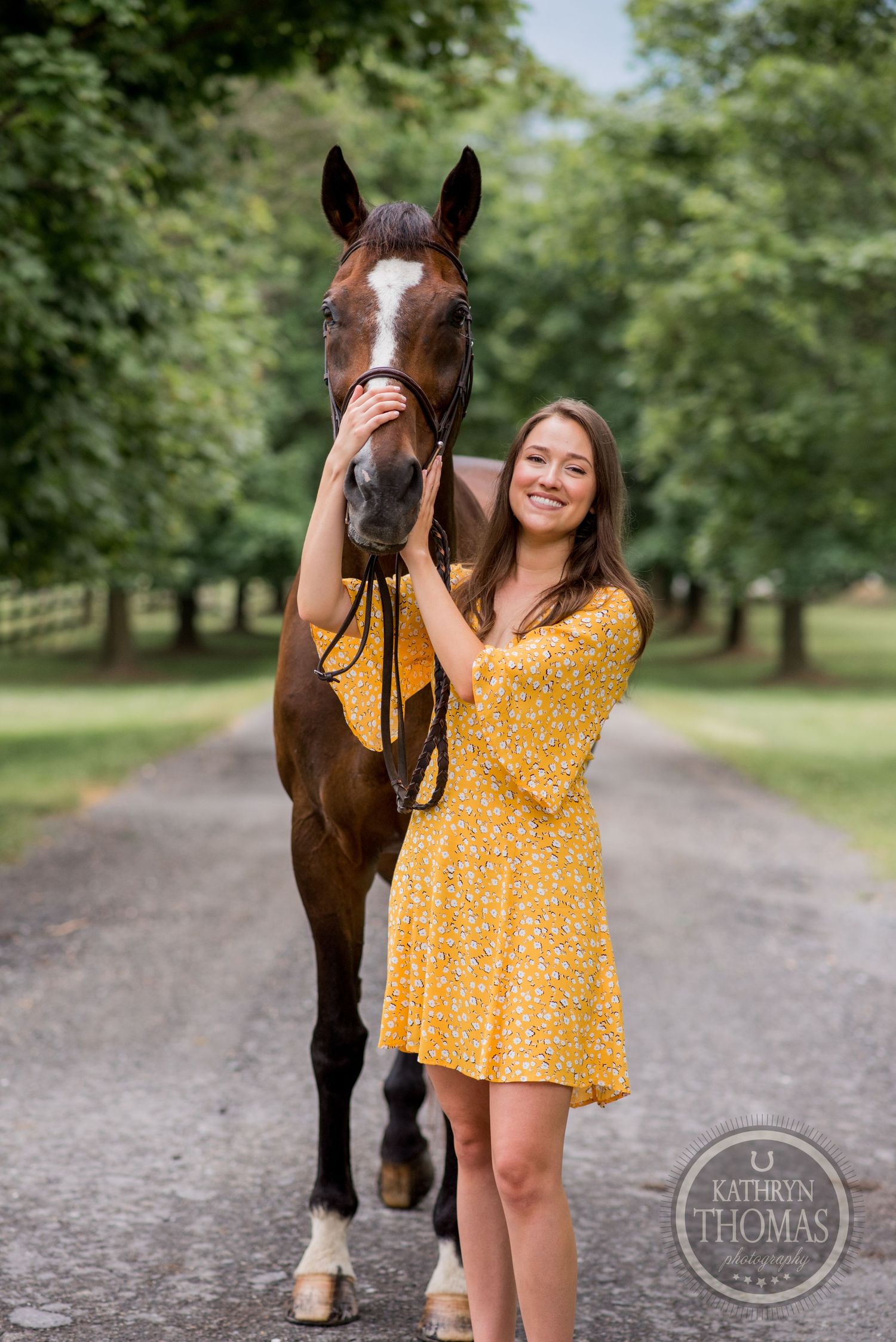 A beautiful bay horse poses with companion wearing a sunny yellow dress on a tree-lined path.