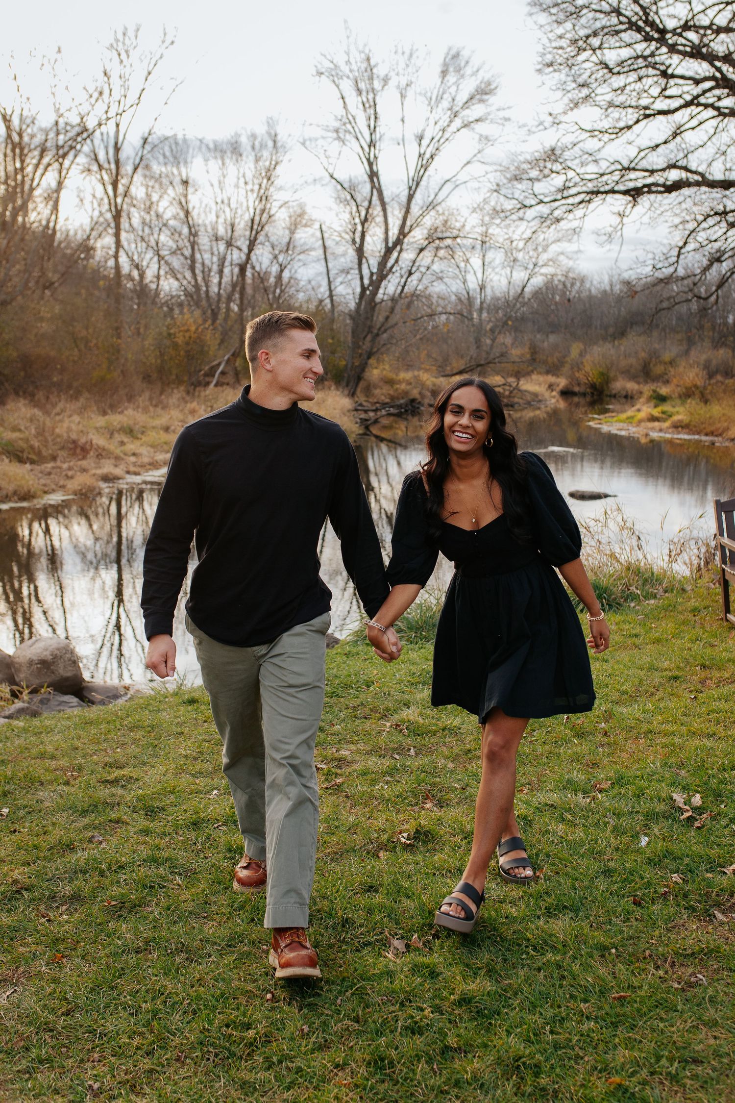 Playful couple running and laughing together on grassy riverbank during autumn afternoon.
