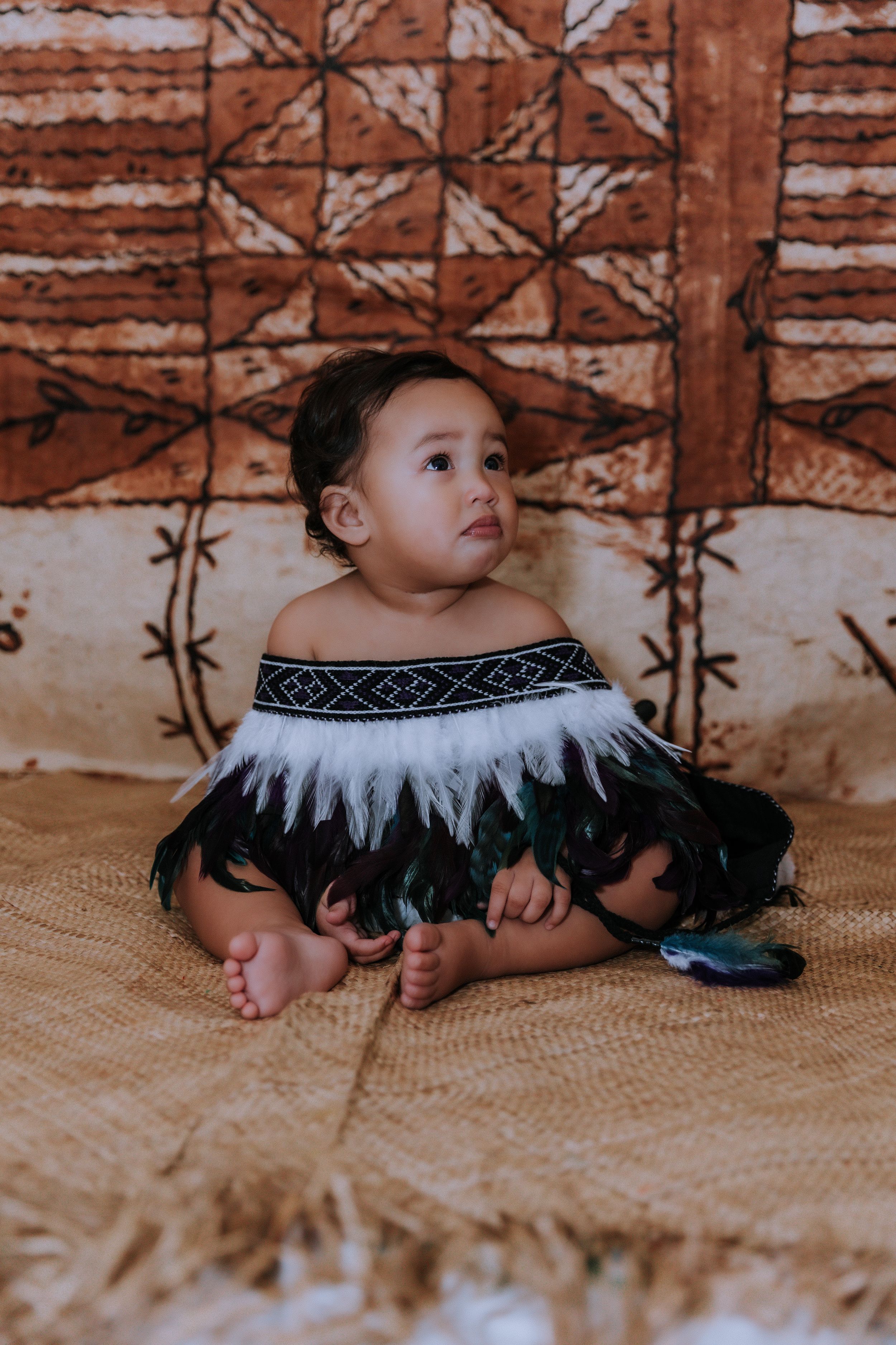 Baby in traditional feathered costume crawling on textured mat against patterned wall.