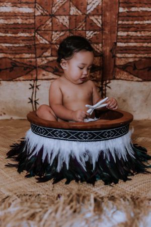 A small child sits in a decorative round basket with black and white feathers against a brown patterned background.