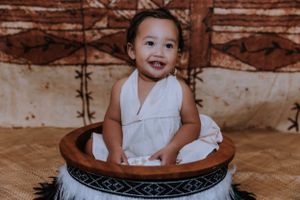 A small child in white clothing sits in a decorated round basket against a brown tapestry background.