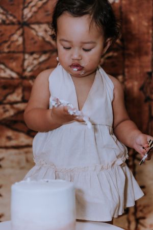 A small child in a white dress reaches toward a white cake on a wooden stand.