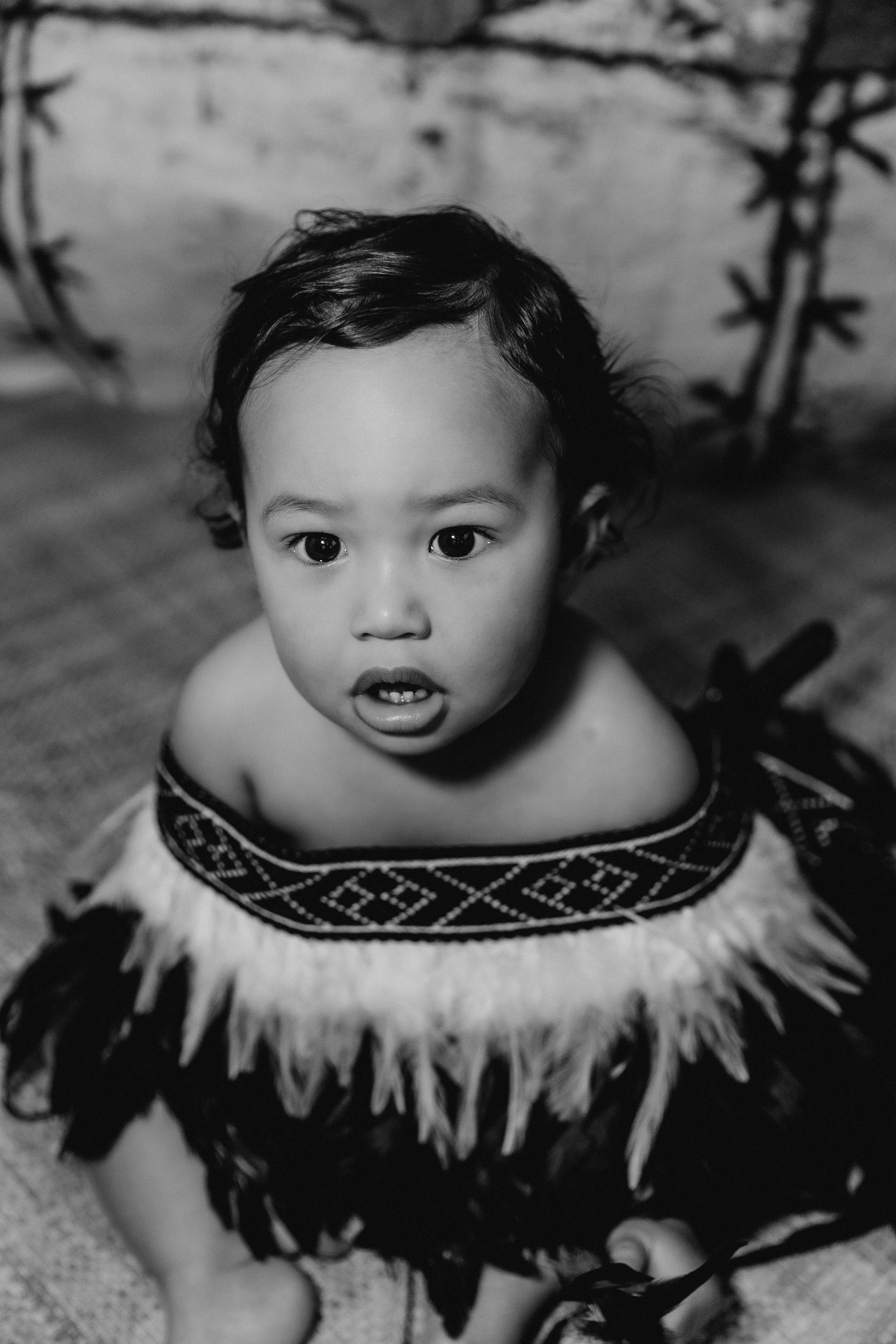 Close-up portrait of baby wearing traditional feathered collar against cultural backdrop.