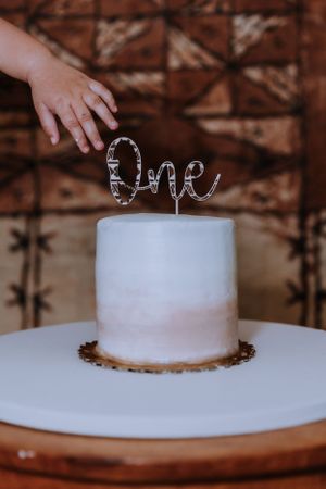 A white cake with silver text topper sits on a wooden cake stand against a brown background.