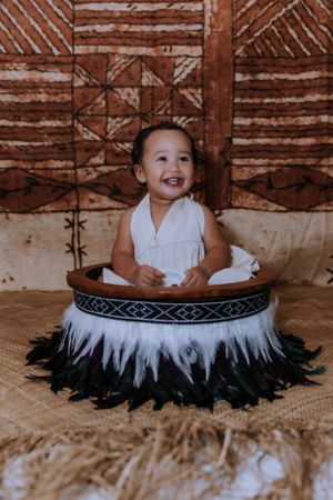 A happy baby wearing white sits in a circular basket decorated with black and white feathers.