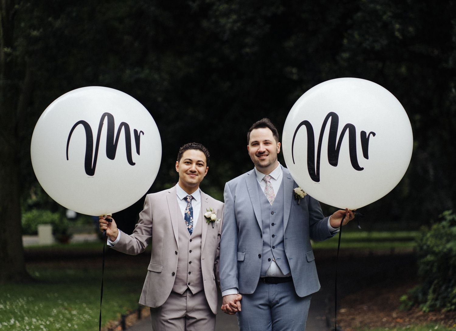 Two grooms holding large white balloons with 'Mr' text in an outdoor setting.
