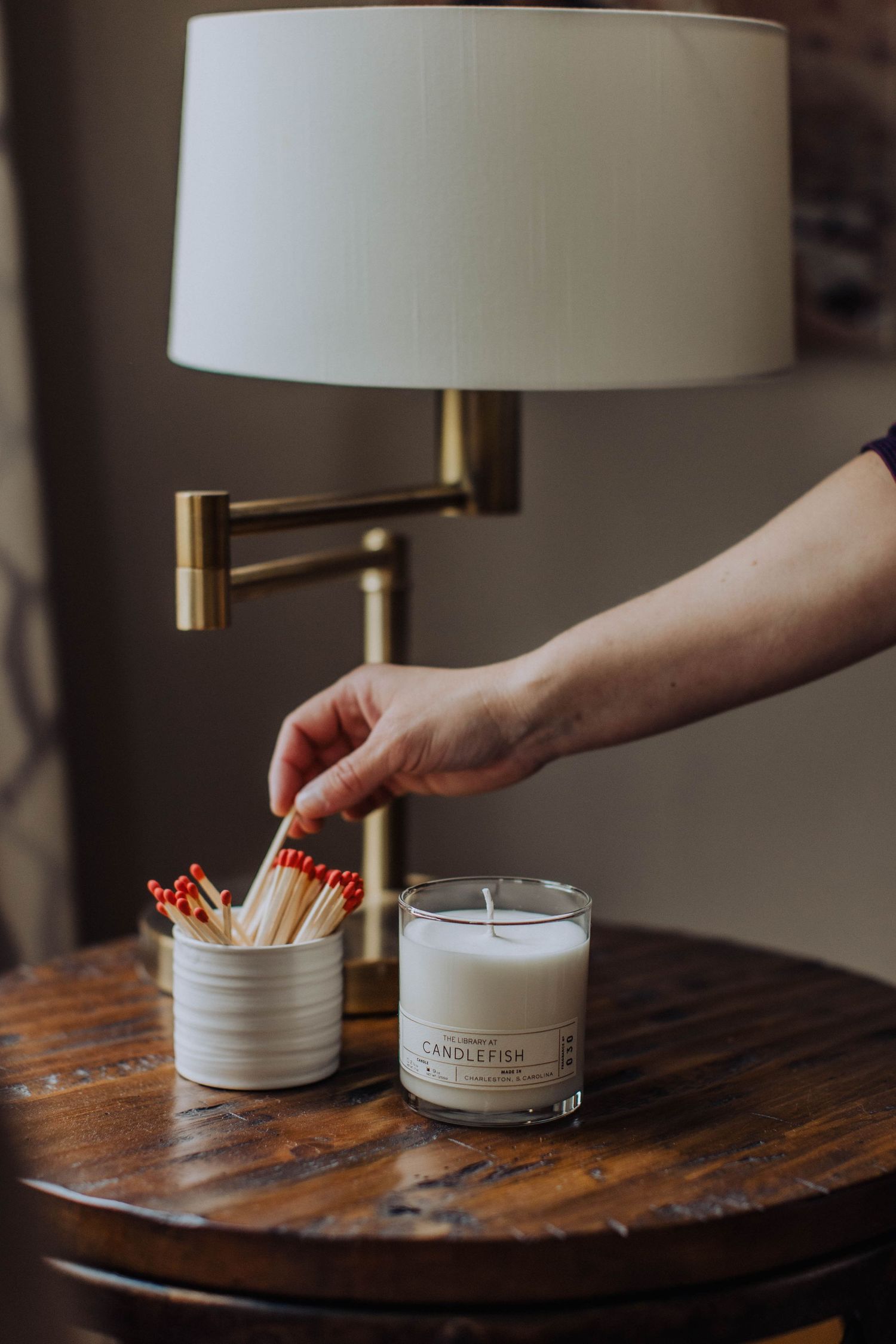 Hand reaching for matches next to white candle and lamp on wooden side table.