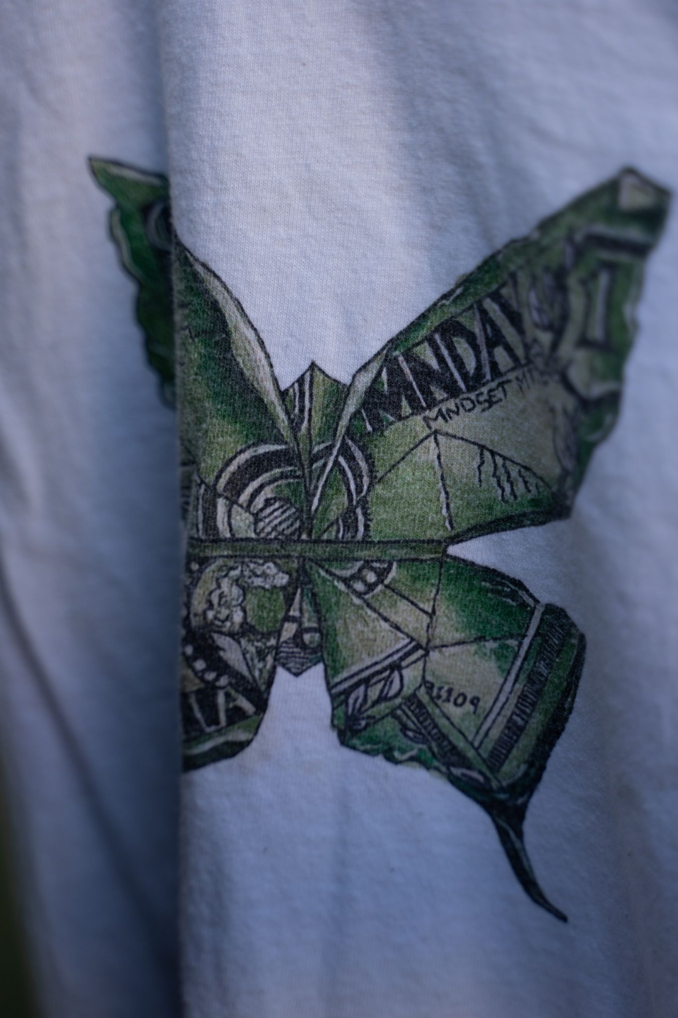 Close-up of a white t-shirt featuring a butterfly graphic made from green dollar bill origami.
