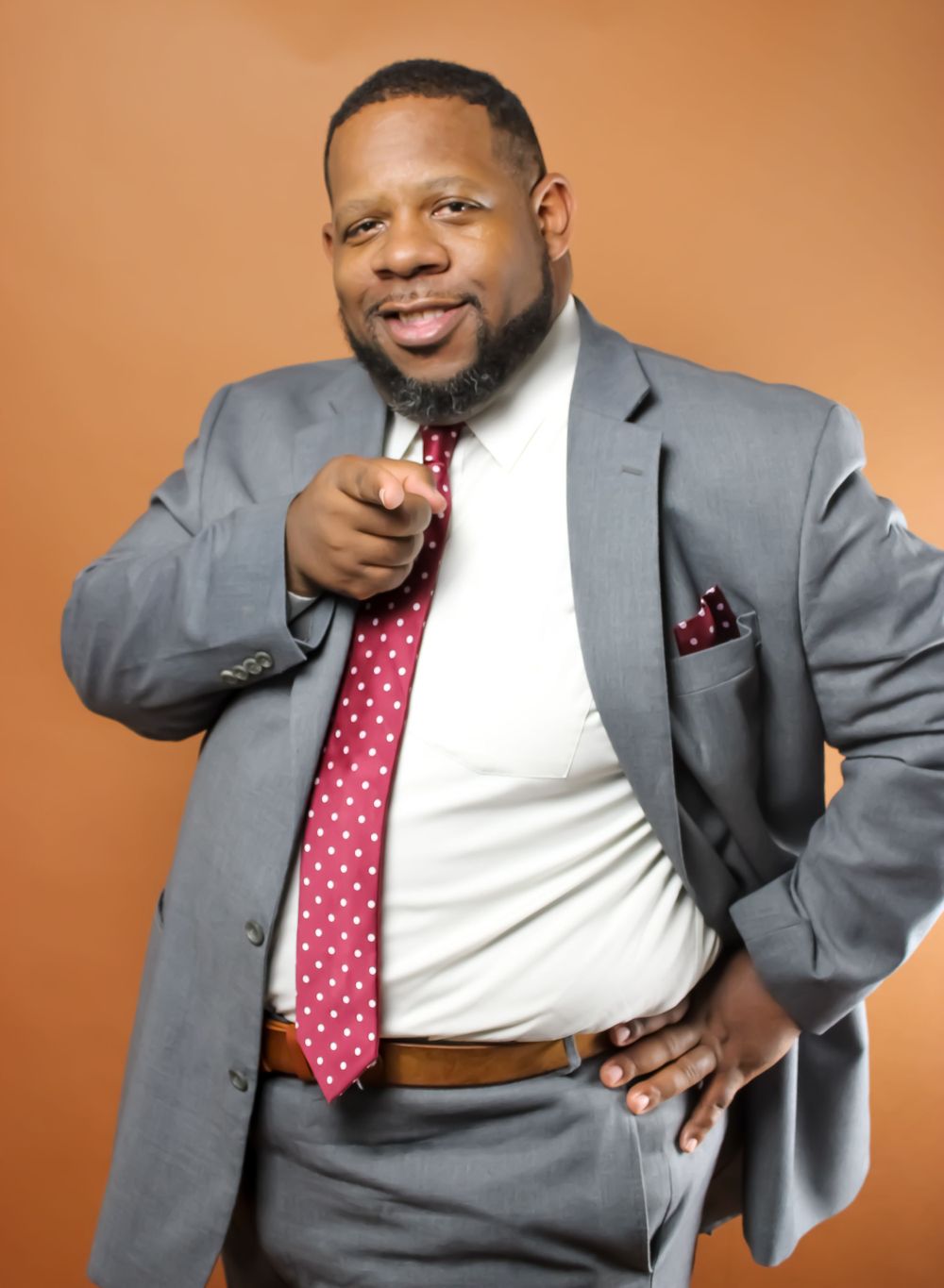 Professional portrait of someone in a gray suit with a red polka dot tie pointing at the camera against an orange background.