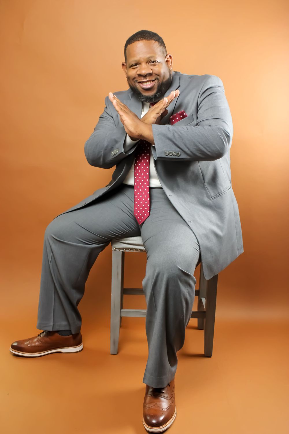 Person in business attire seated on a stool making a crossed arm gesture against an orange backdrop.