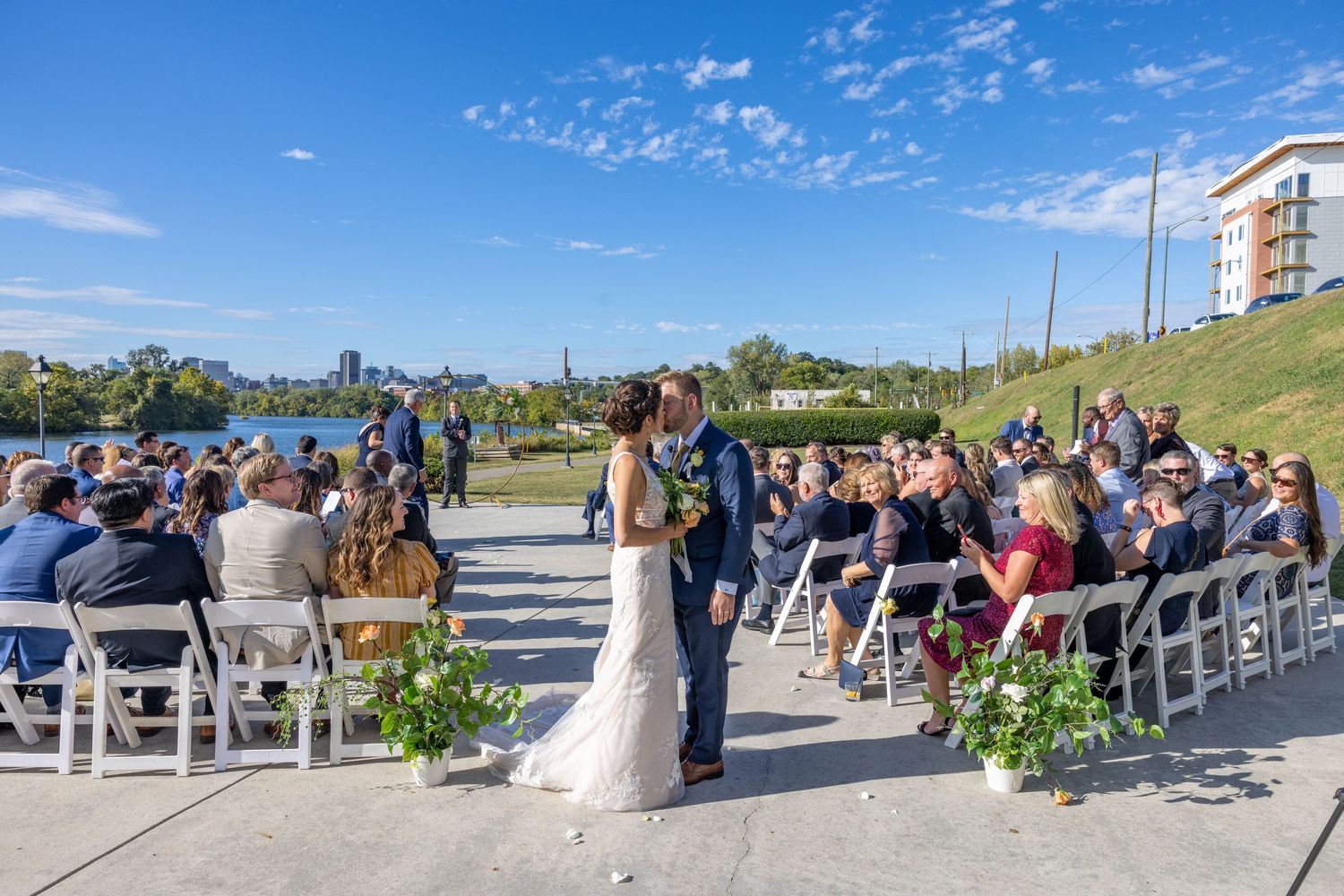 Outdoor wedding ceremony setup with white chairs and guests seated under a bright blue sky.